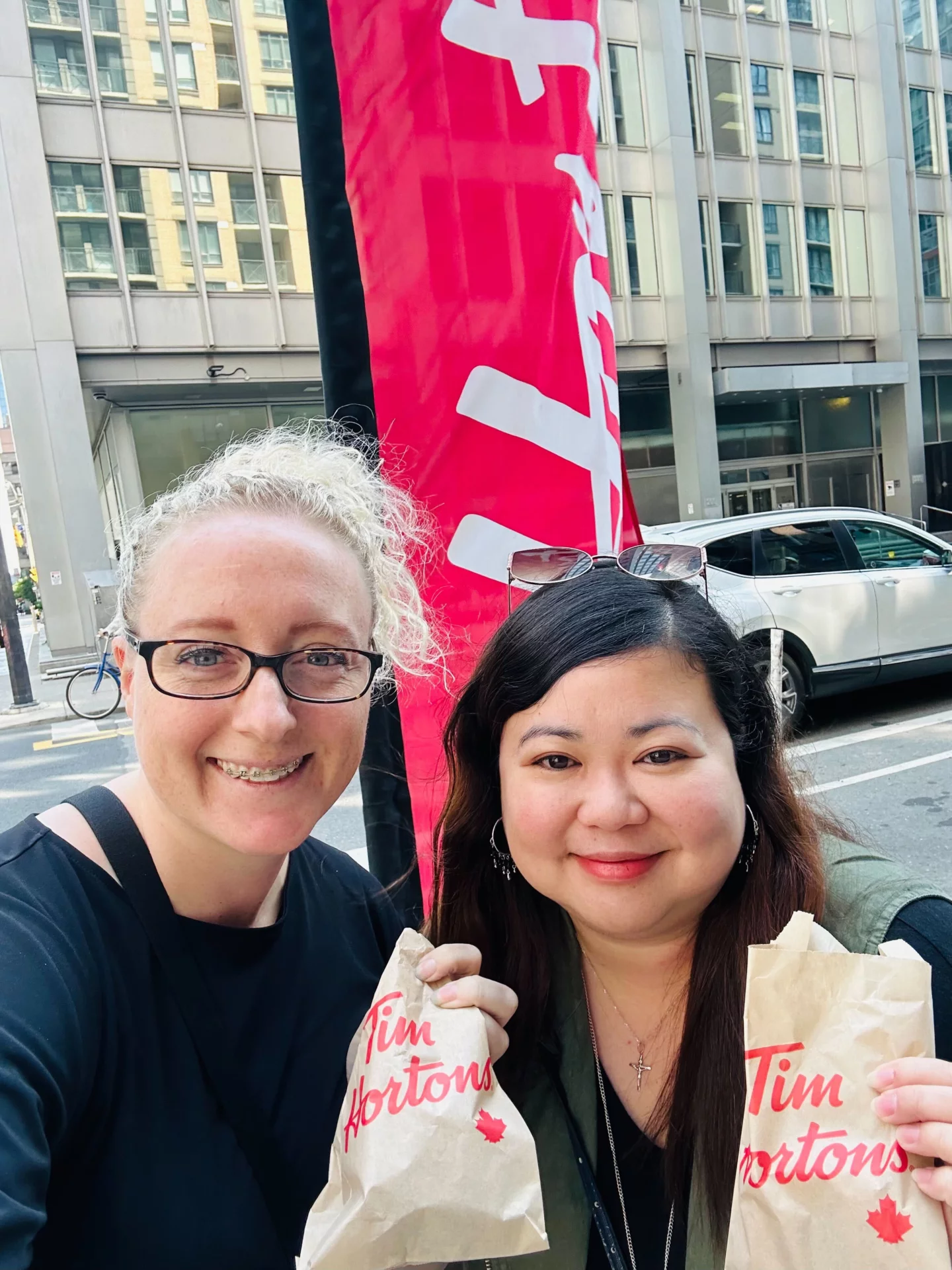 Jackie and Maria are smiling at the camera outside a cafe in Toronto. They have takeaway food (maybe donuts). Maria is visiting from the Philippines.