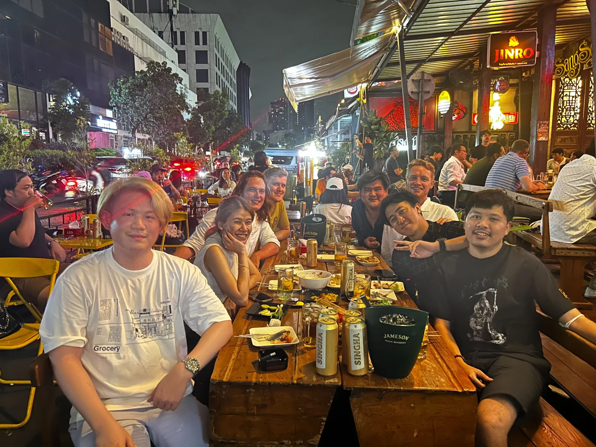 Some of the Bangkok team smile at the camera whilst having dinner and drinks outside a restaurant in a busy street . Daniel and Tom are visiting from Cape Town.