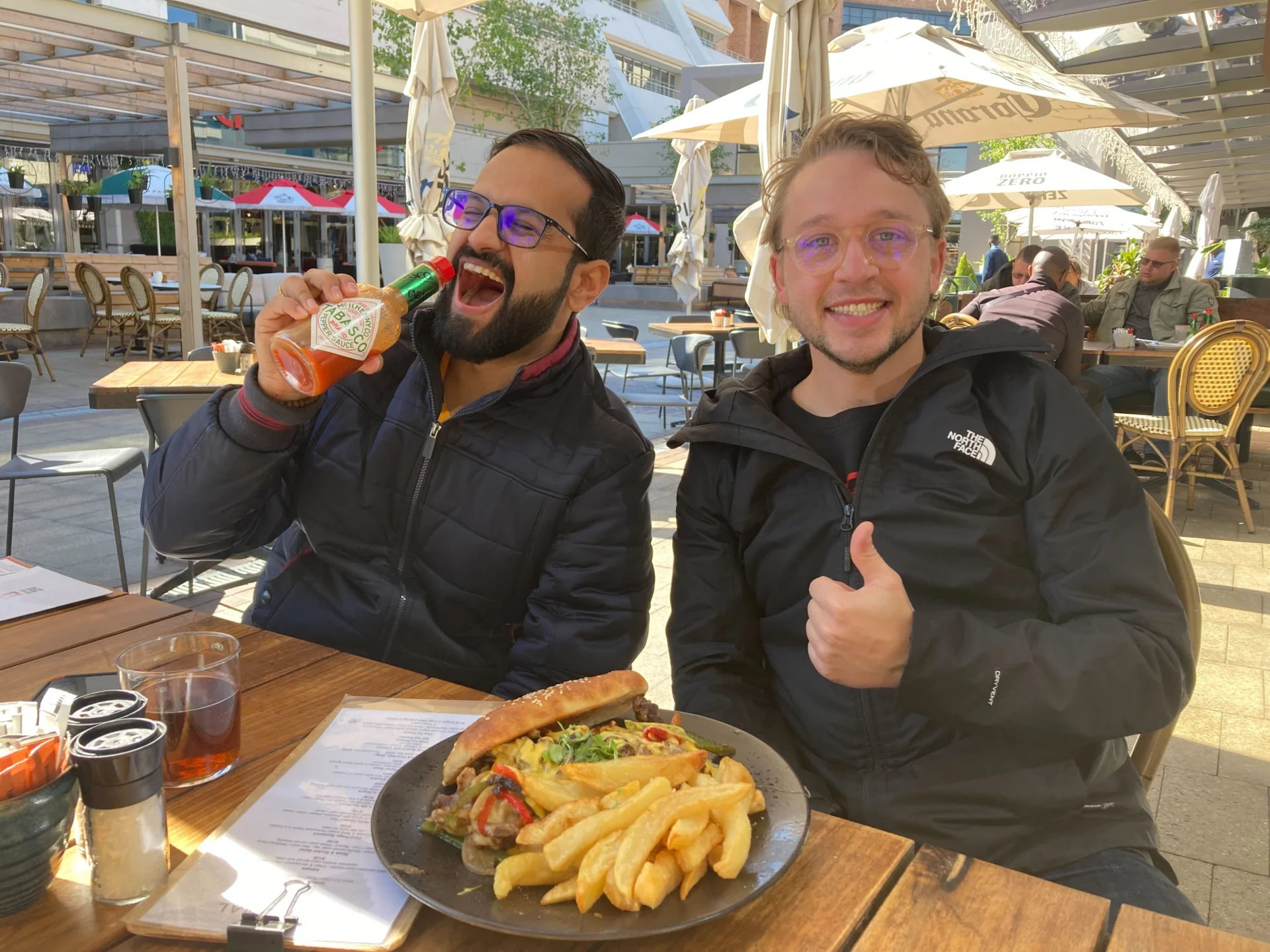 Jason and Yeeshu enjoying lunch during a break from project work. The is a big plate of sandwich and chips on the table, Jason looks very happy about it.